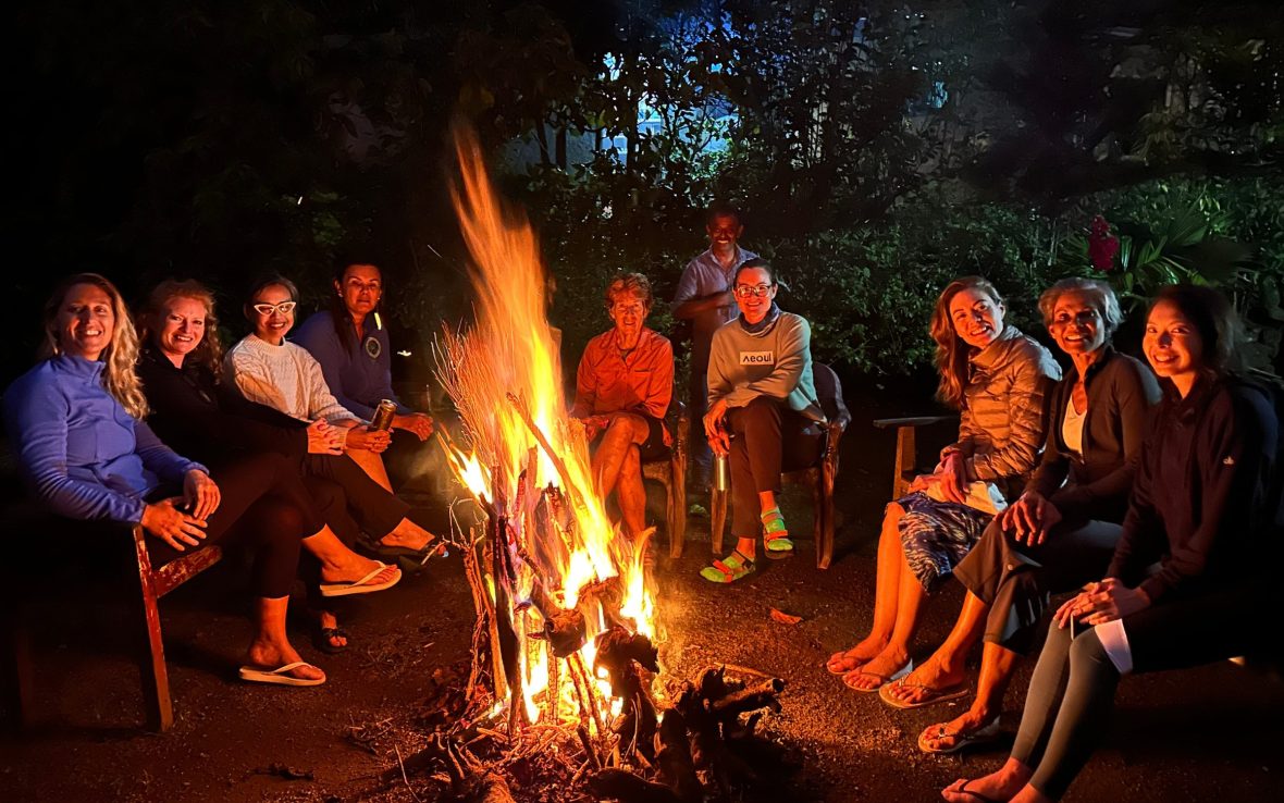 A group of women sit in a semi-circle around a campfire, smiling.