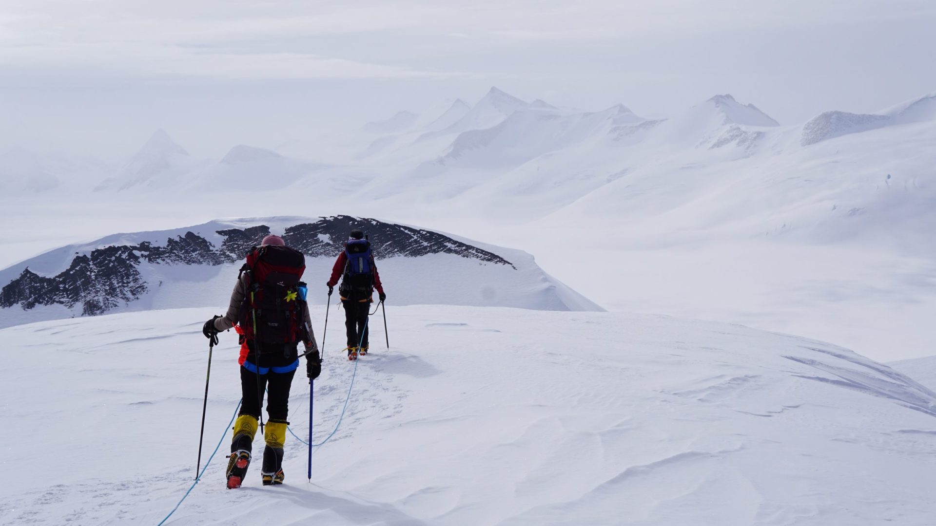 Two women cross-country ski across a snowy mountainscape.