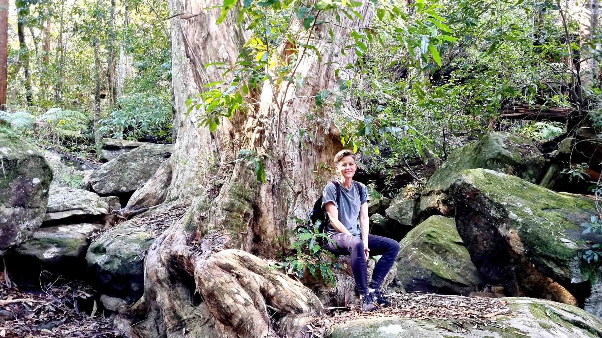 A person sits by a tree and smiles to camera.