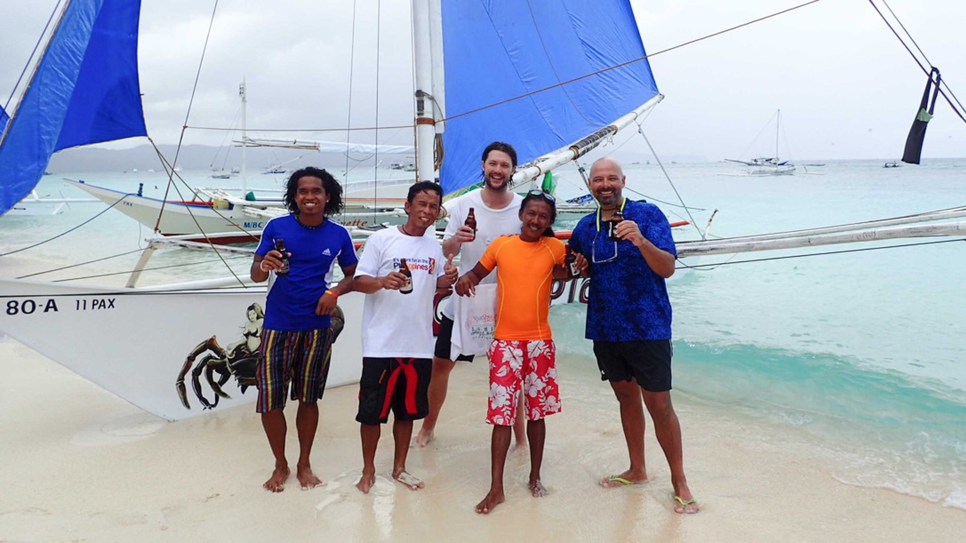 Several men smile with beers, in front of a sail boat.