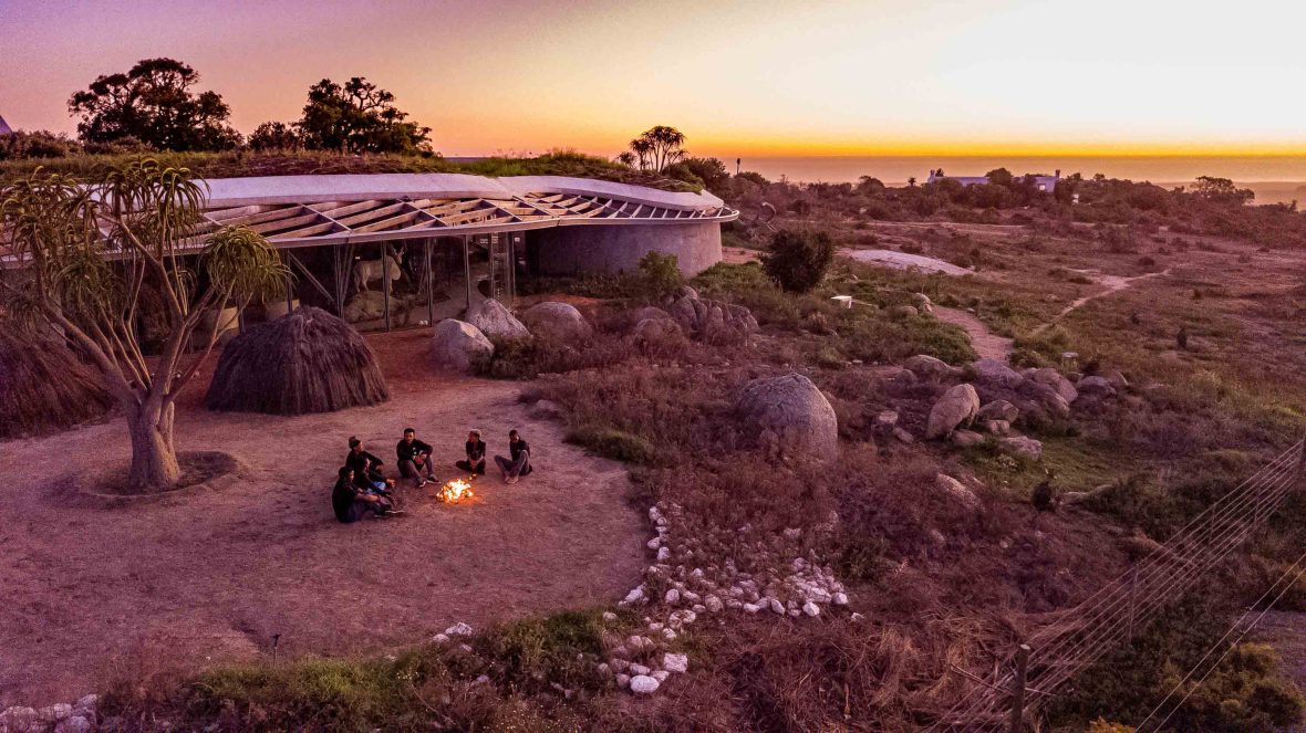 People sit around a camp fire in front of a large building at sunset.