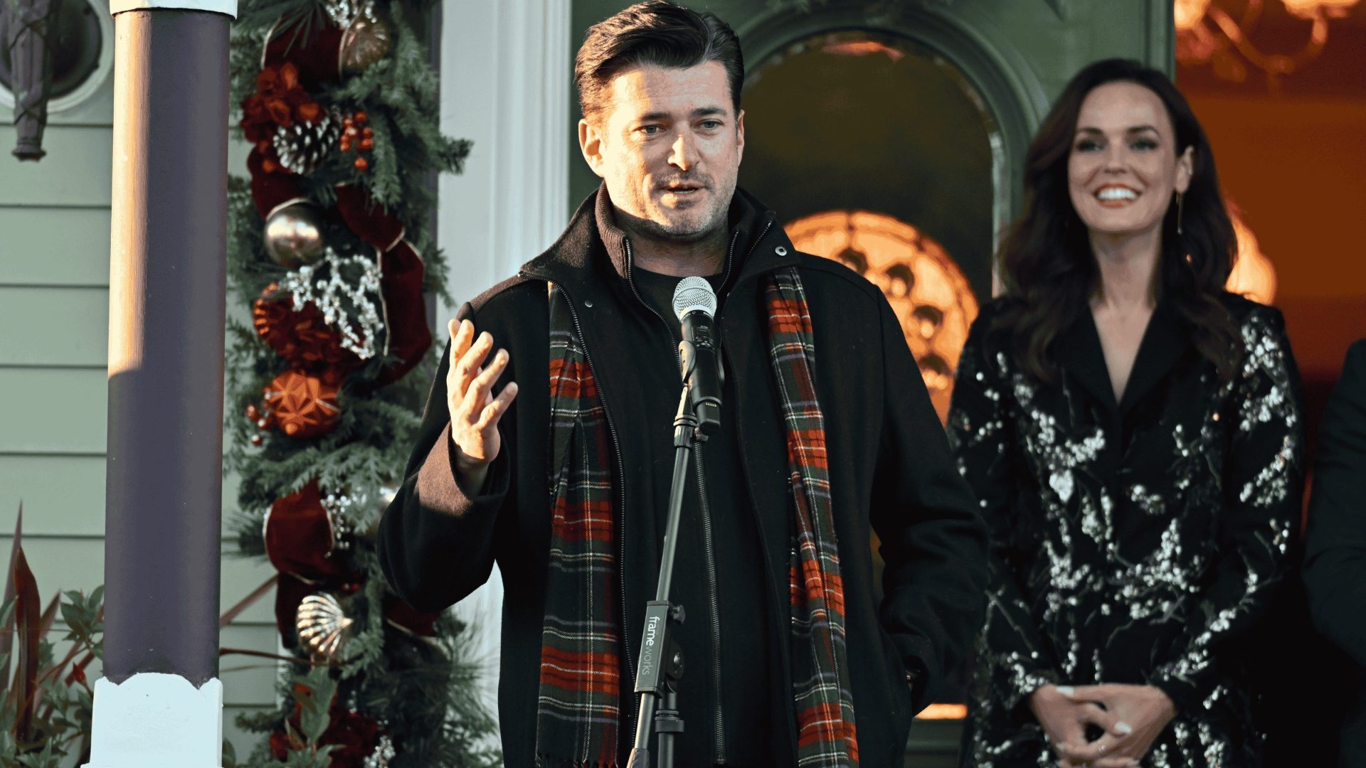 A man in a black jacket and plaid scarf gives a speech next to a Christmas tree. A woman stands to the right.