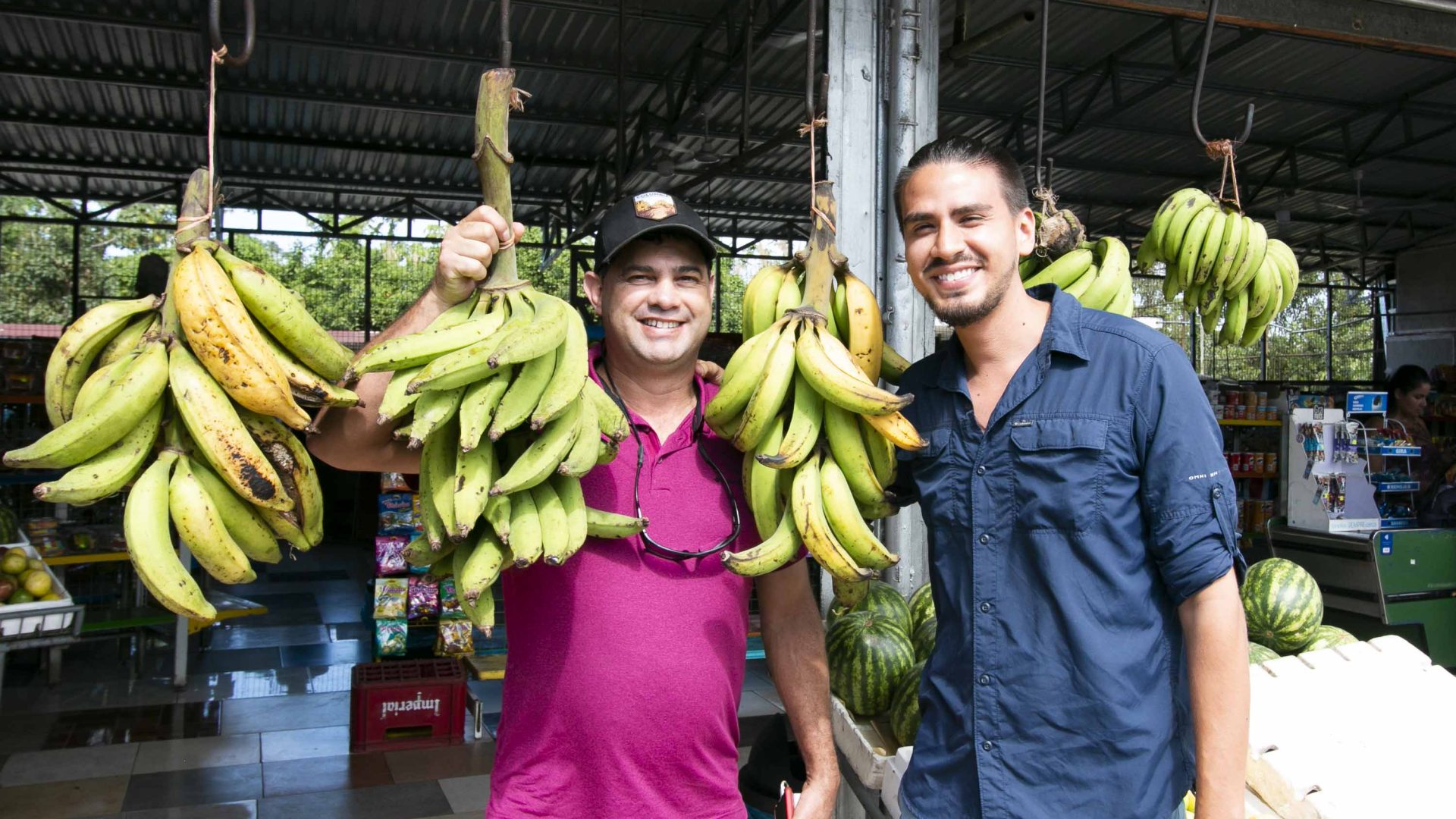 Two men hold up bunches of bananas at a fruit stand called Fruteria Krystal.