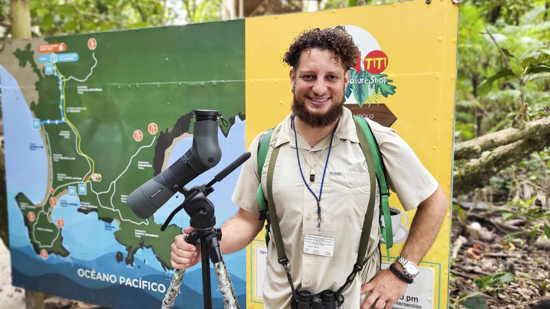 A tour guide stands in front of a sign for the National Park. He holds a telescope.