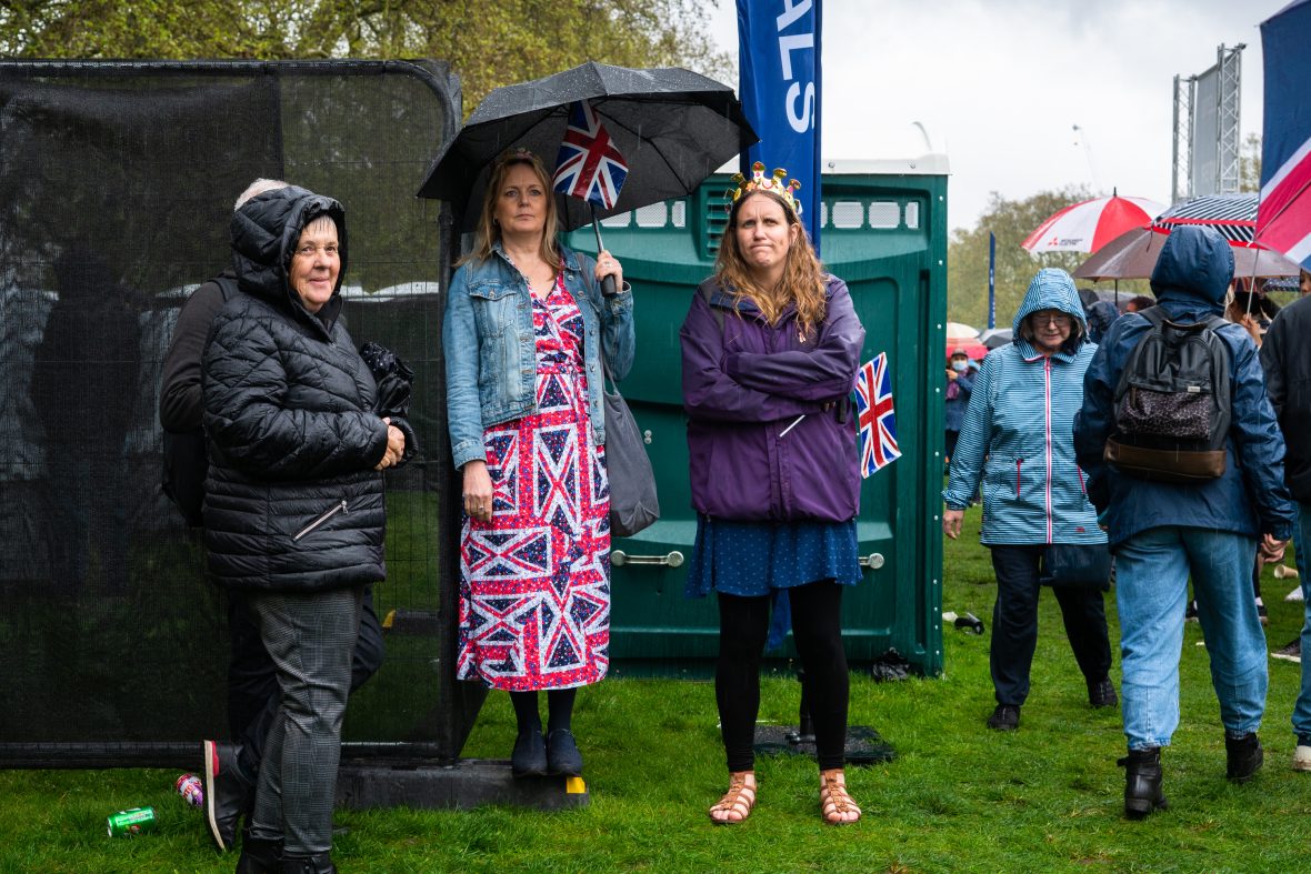 Three women posing for a photo; the middle one wearing a Union Jack dress and holding a flag, while one wears a crown.