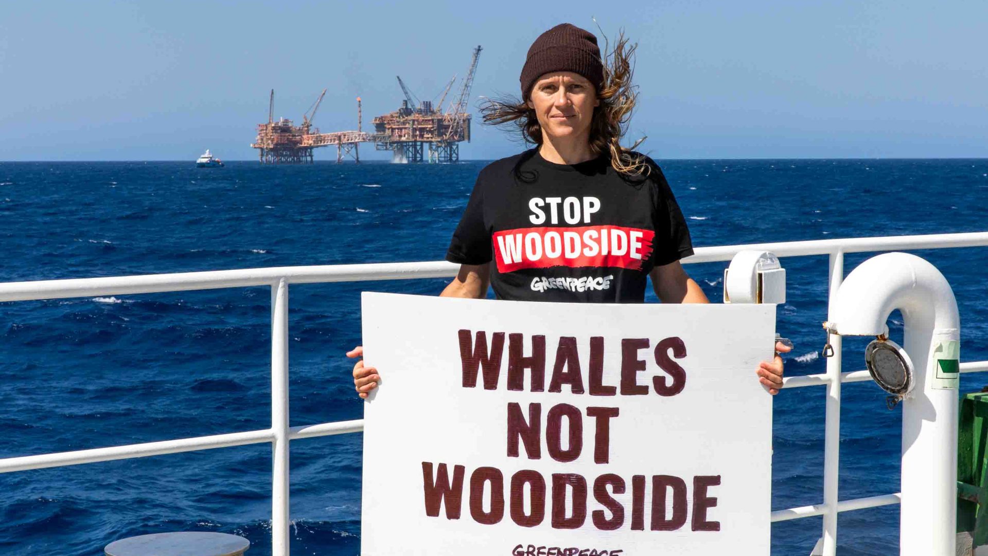 A woman stands on a boat with a serious expression. She holds a sign that says "Whales not Woodside". Other boats are in the background.