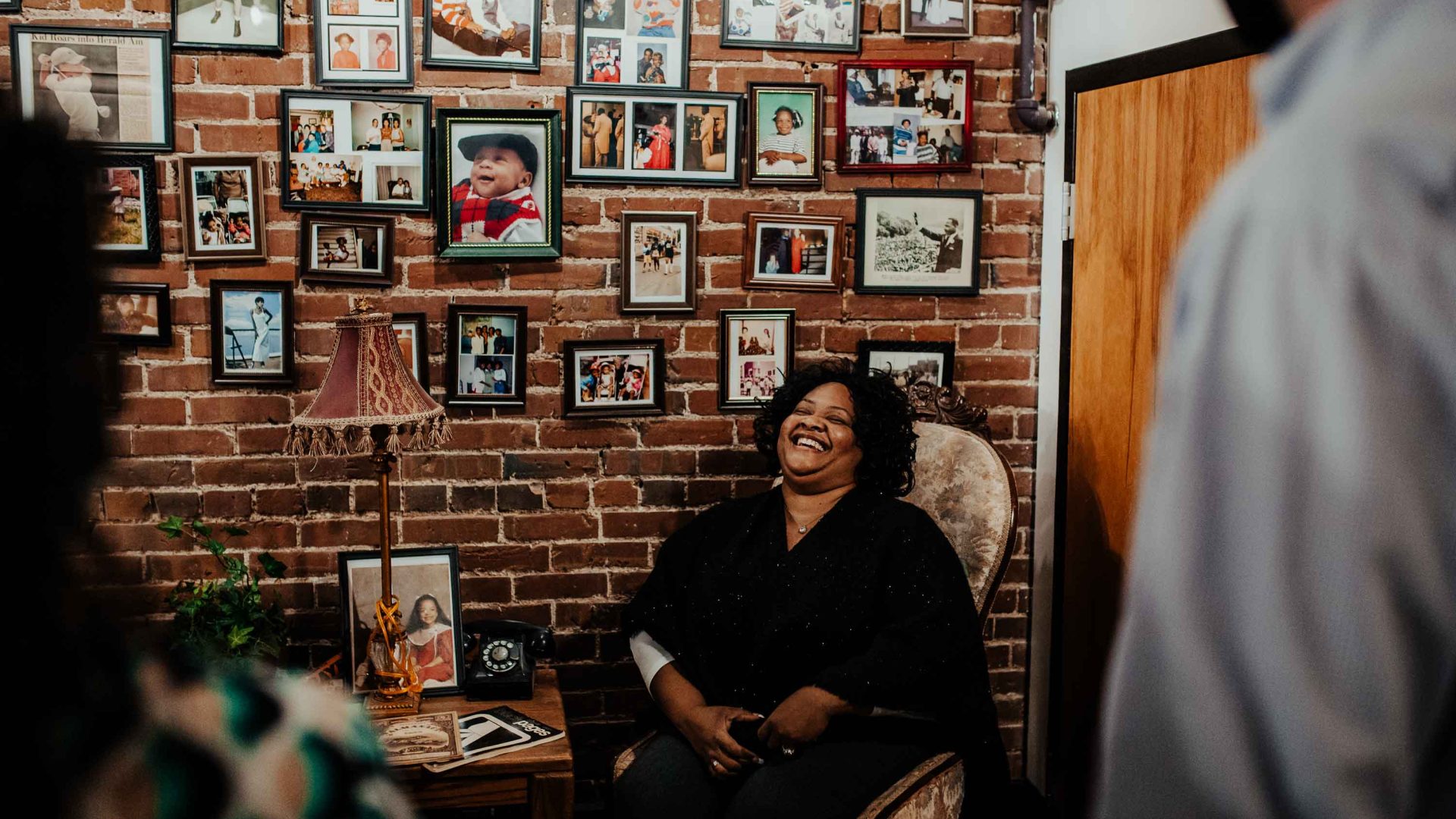 A woman sits on a chair at the museum and laughs. She's next to a wall of photos.