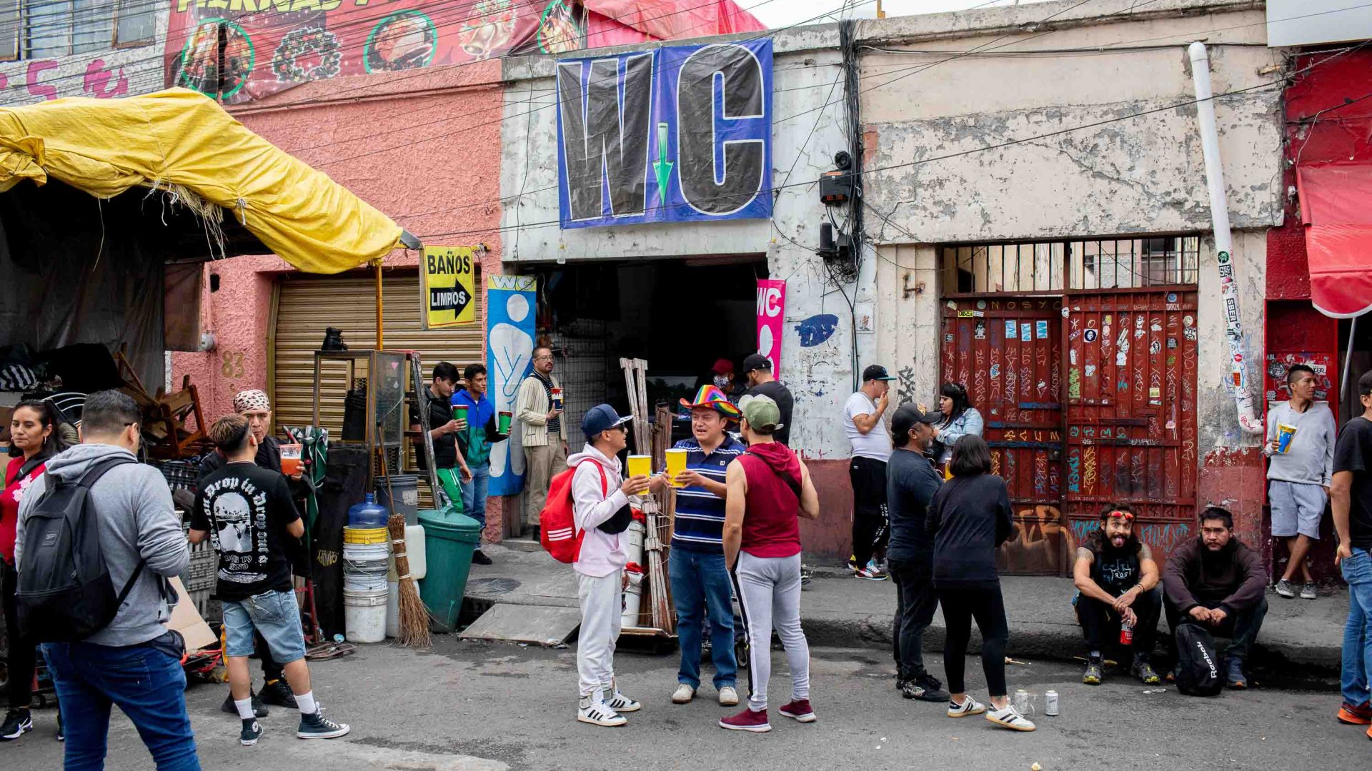 A group of people stand outside the public restrooms drinking micheladas.