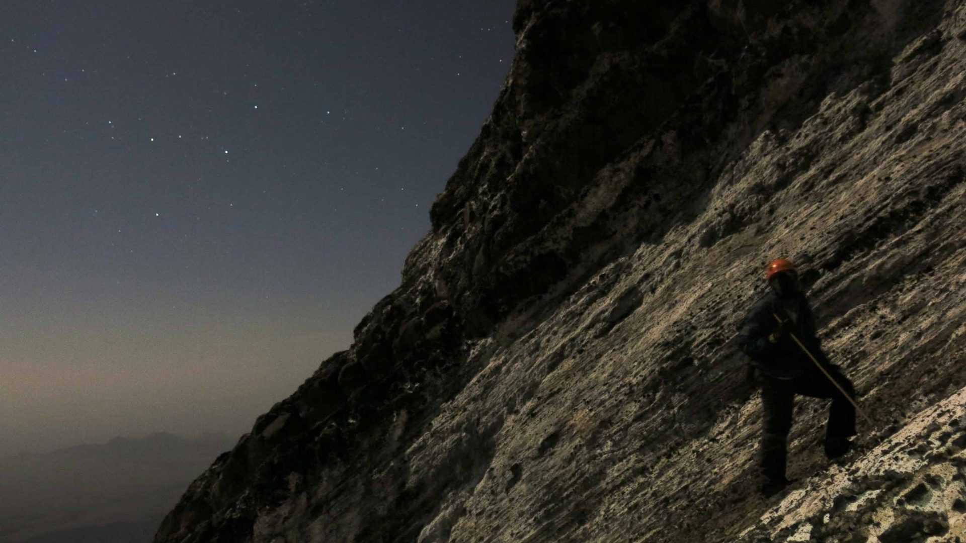 A person is barely visible as they climb a sheer cliff at night.