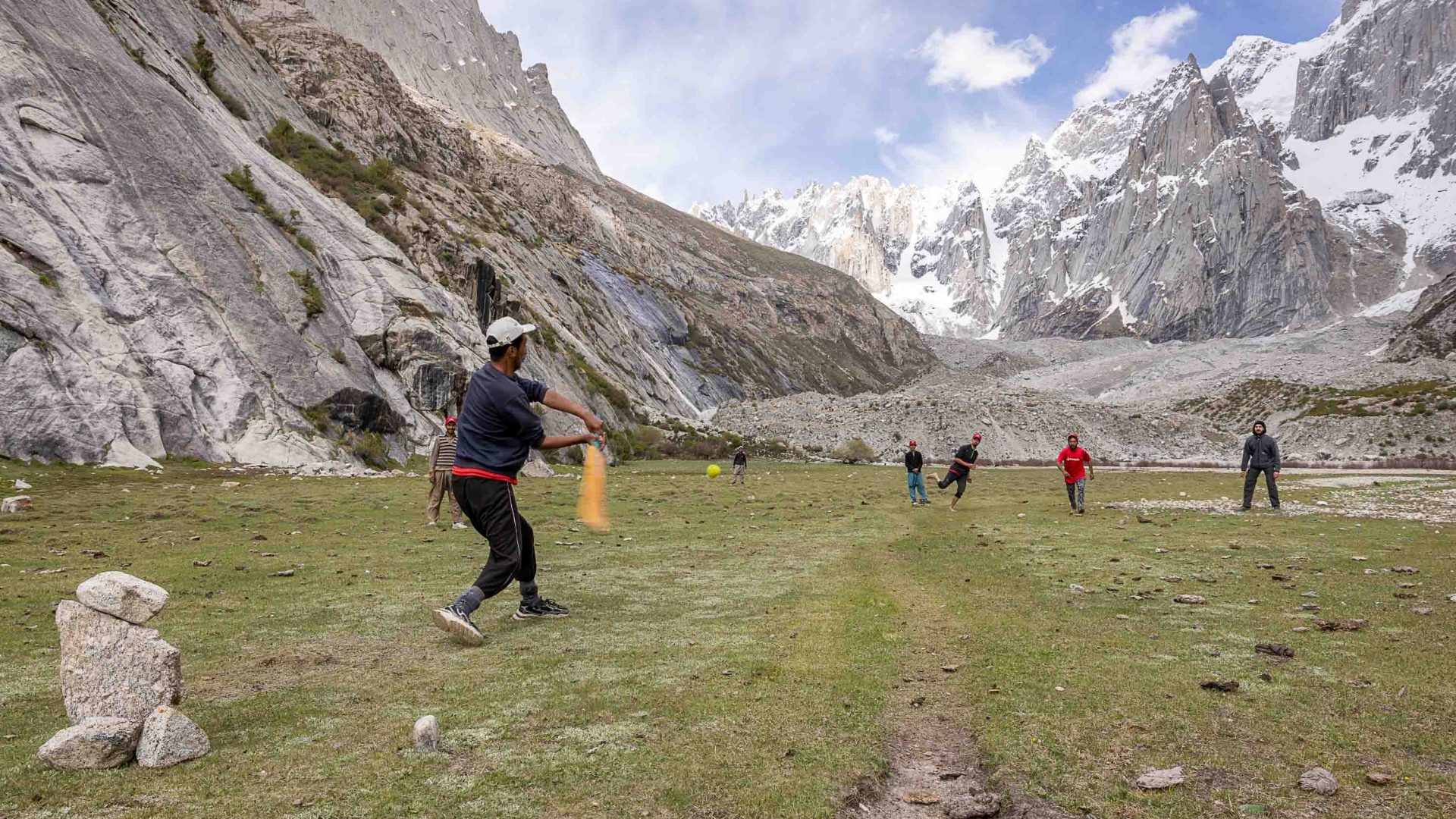 Trekkers and porters play cricket together below mountains.