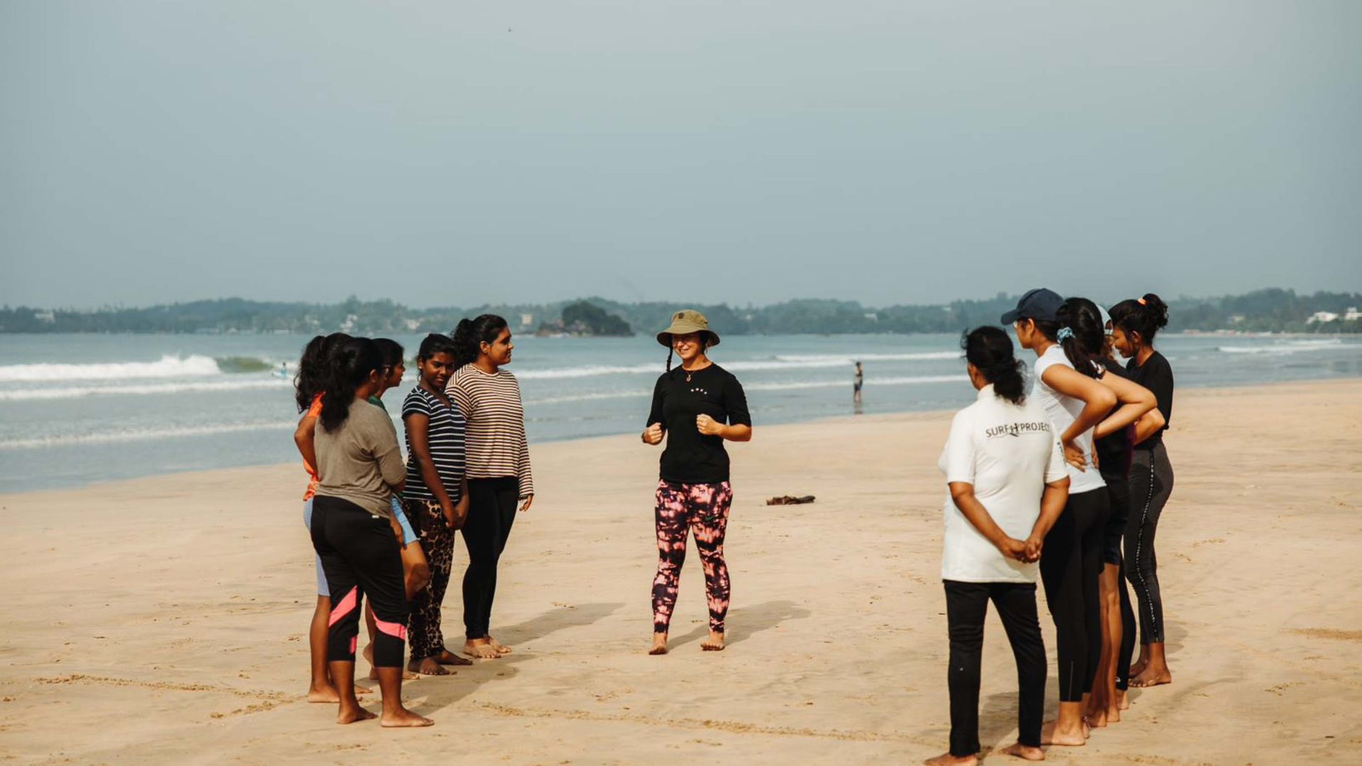 A group by the beach listens to some instructions from a person in a hat.