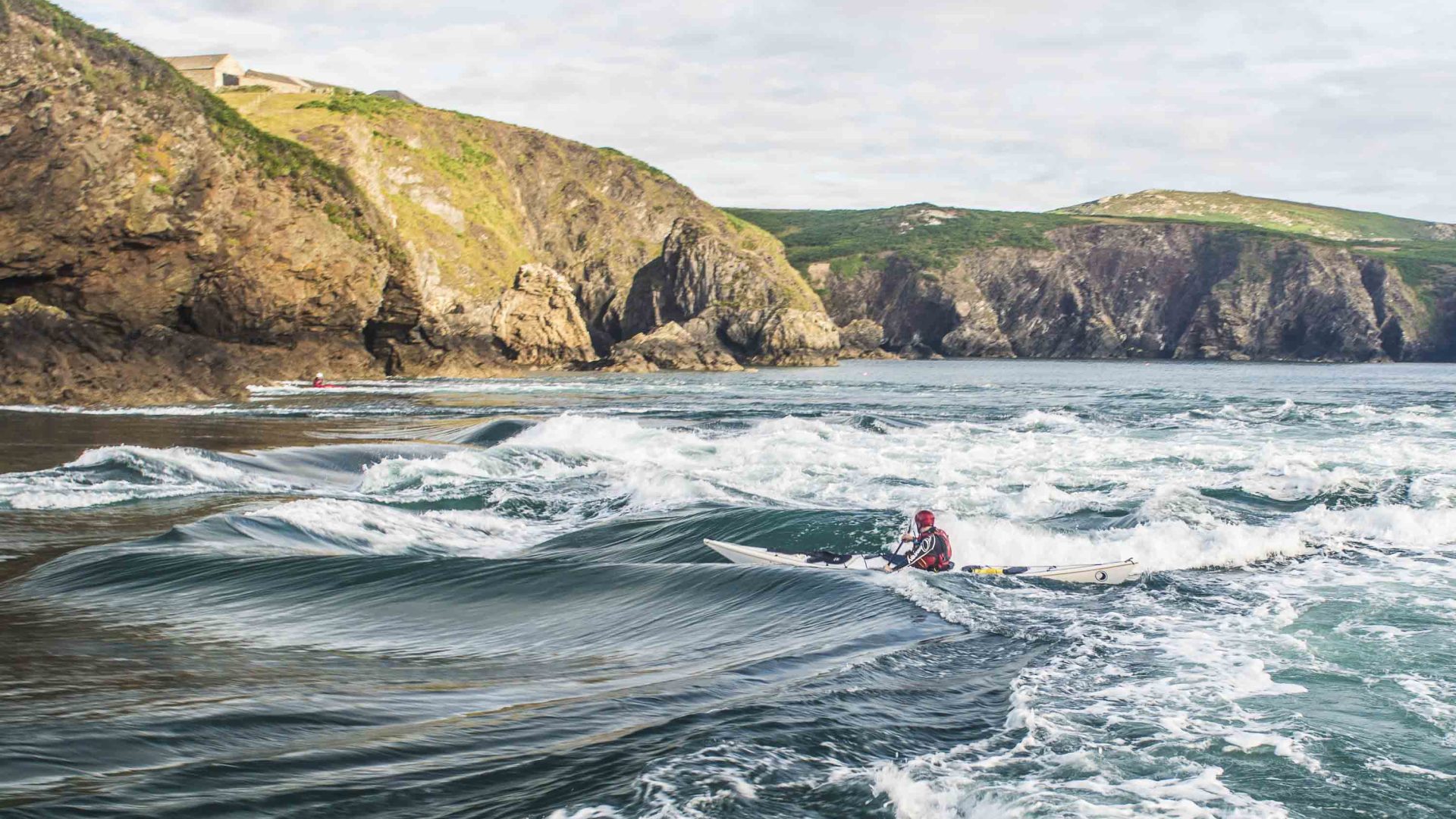 A man kayaks on waves near some rocky cliffs.