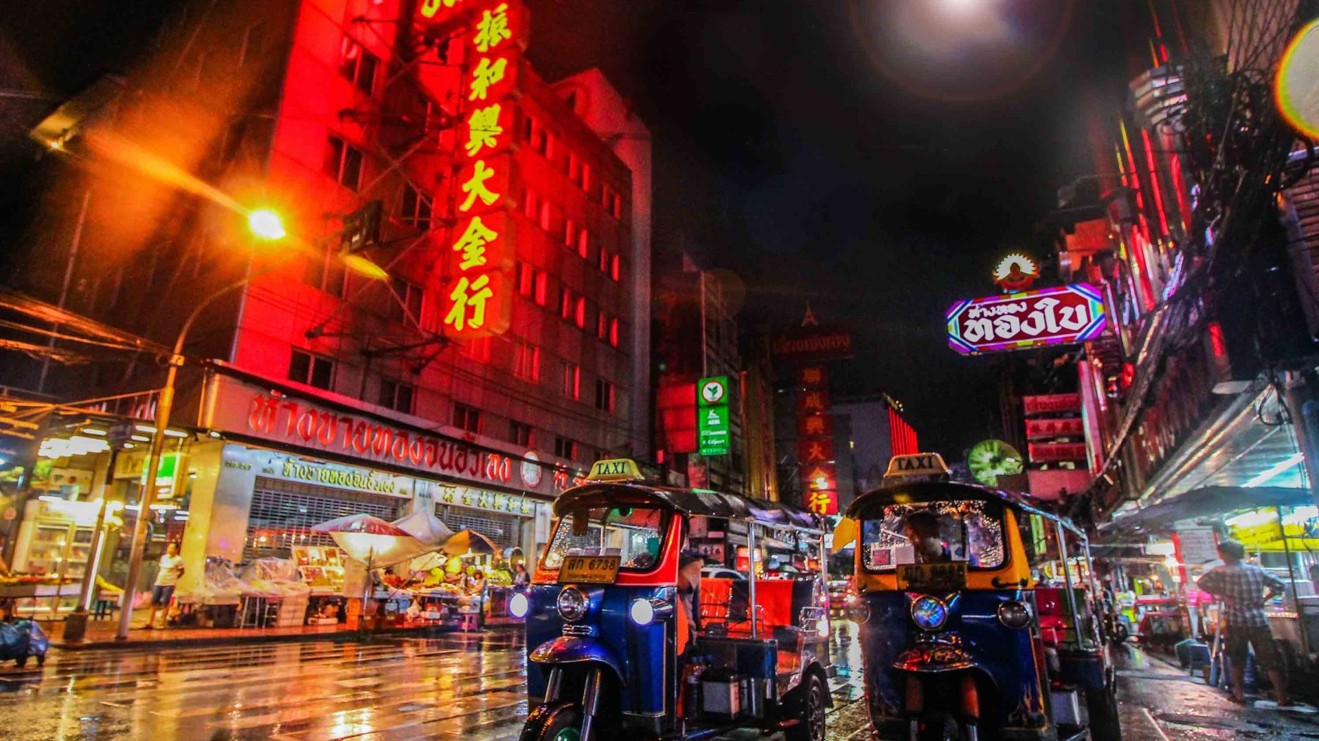 Two tuk tuks in a street at night which is full of bright lights and signs.