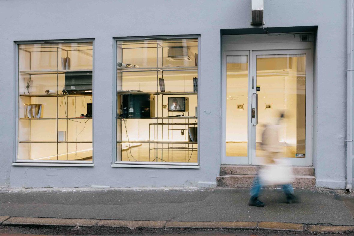 A person walks past the windows to the exterior of the Fotogalleriat in Oslo. The light outside is low, while it is brightly lit up.