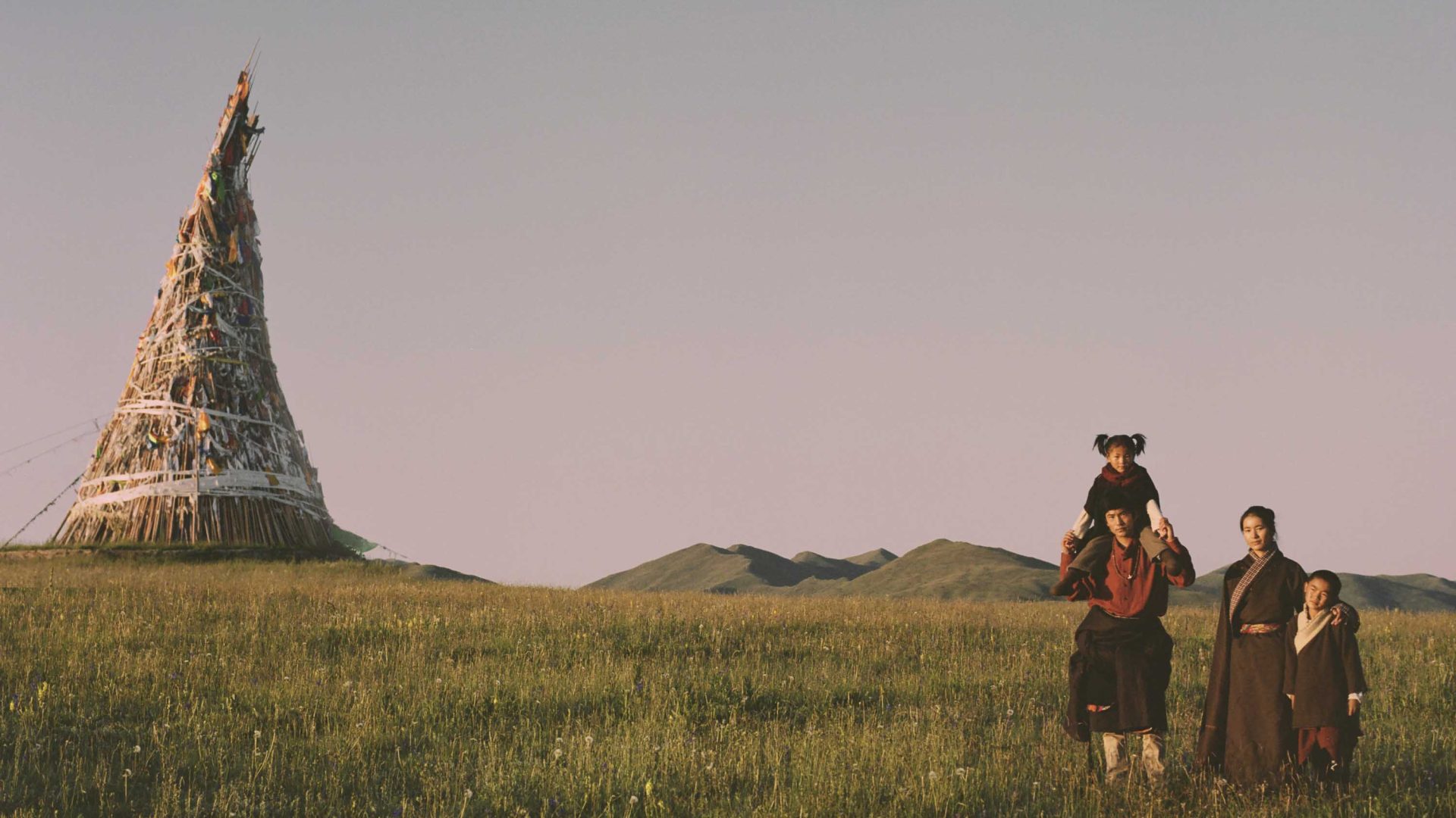 A family walks together through the grass. There is a tipi shaped structure in the background.