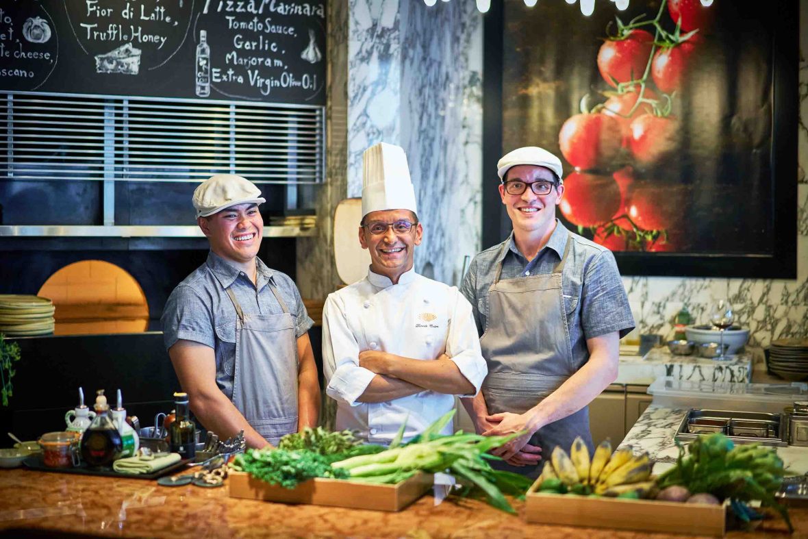 Three chefs smile to camera in front of a table of ingredients.