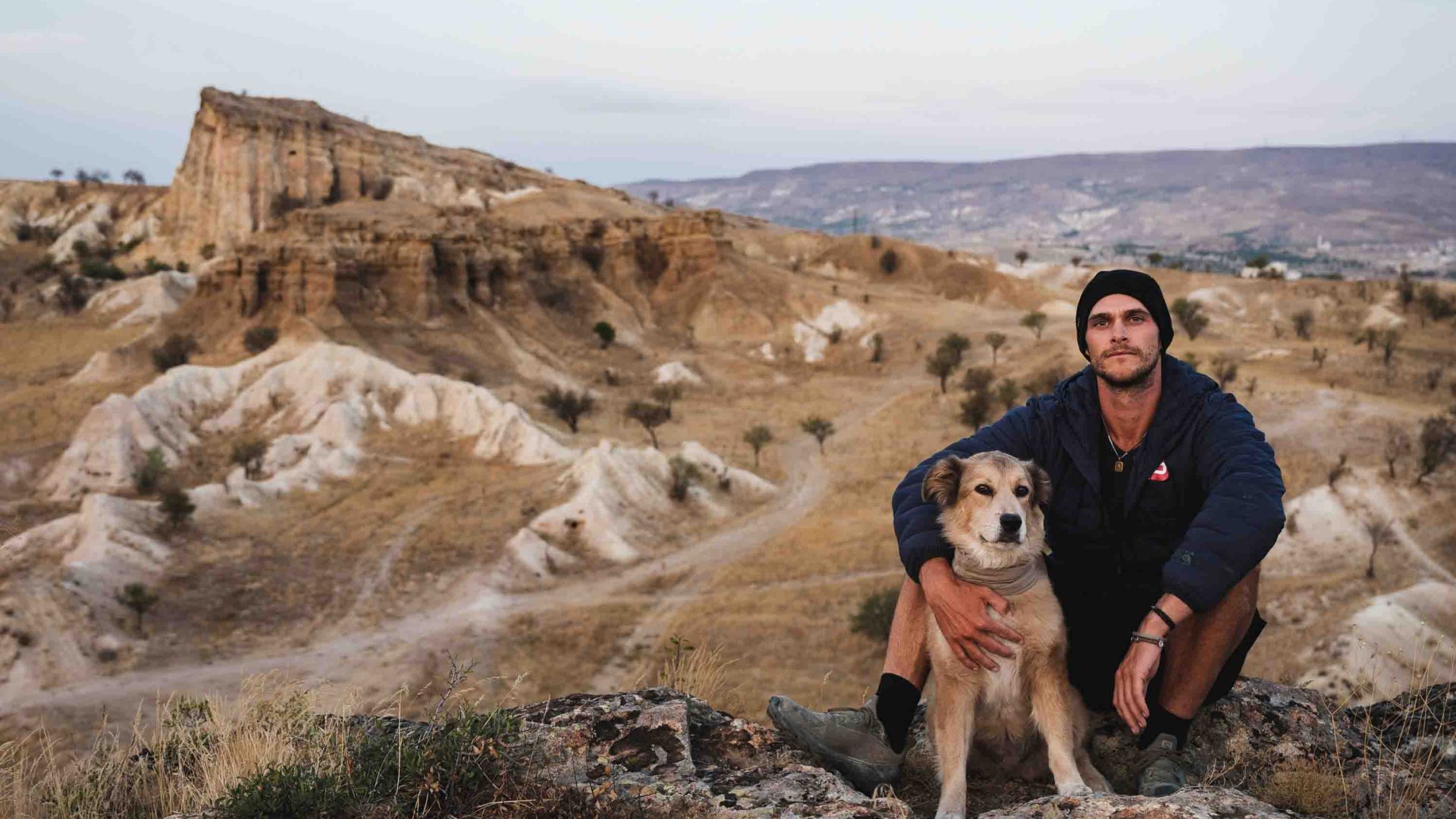 A man and his dog pose for a portrait on the ridge of an arid valley.