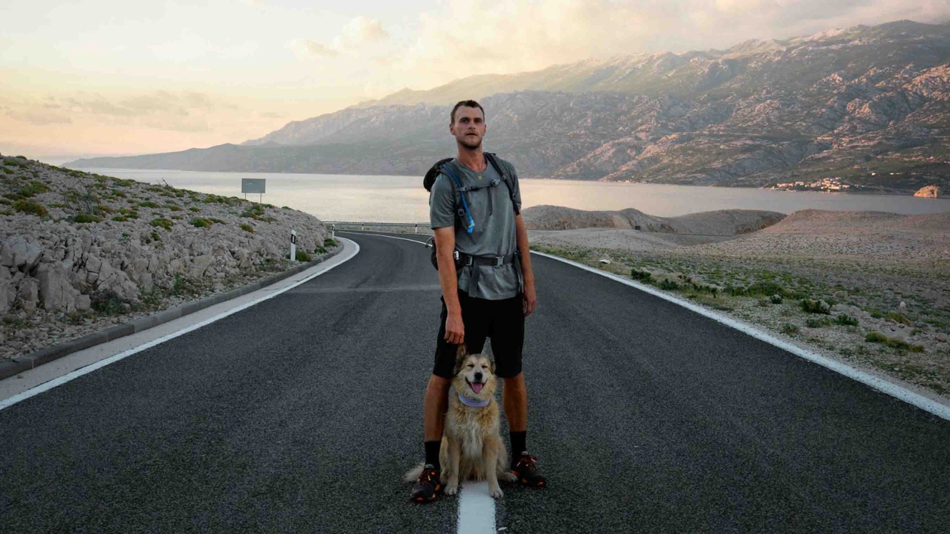 A man stands in the middle of the road with his dog. There is a lake behind him.