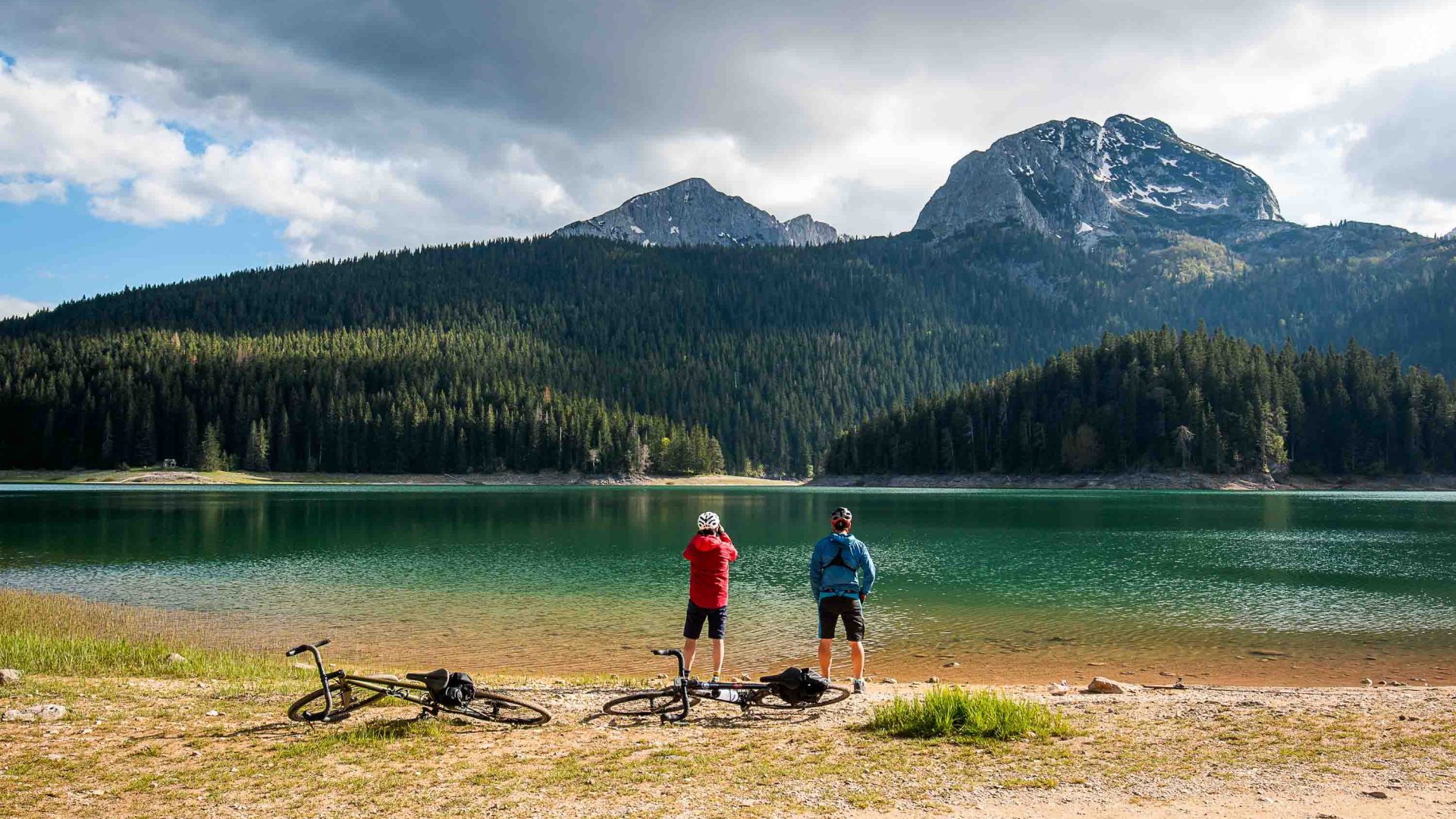 Two cyclists look out at a green lake in front of mountains.