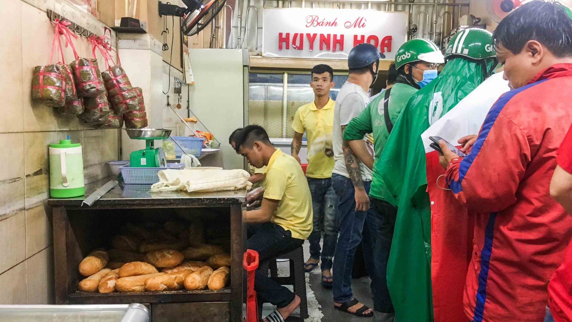 A row of people wait for their bahn mi order while a seated man prepared them.