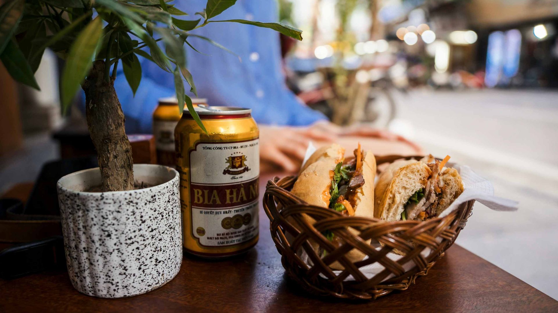 A bahn mi in a basket, on a table with a beer.