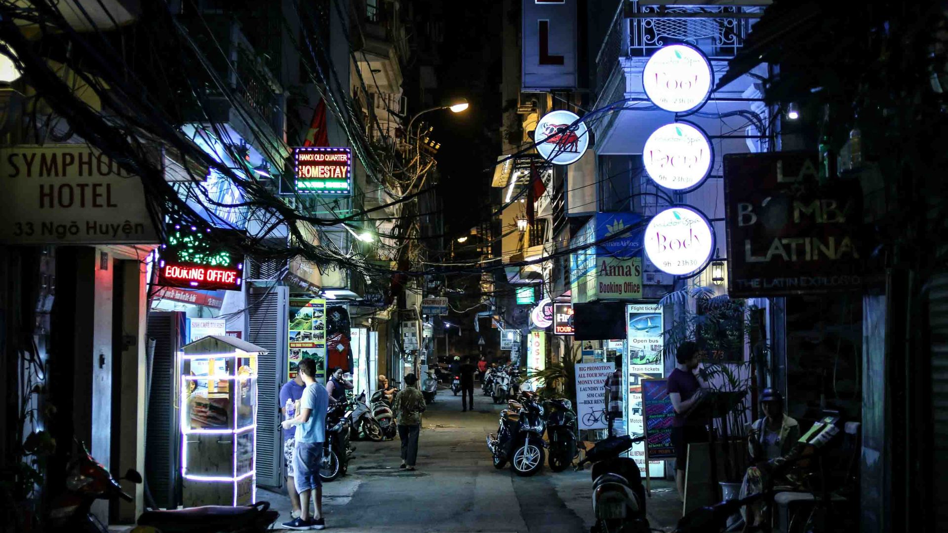 A person stands in the light of a bahn mi stall in a street packed with small businesses in Hanoi at night.