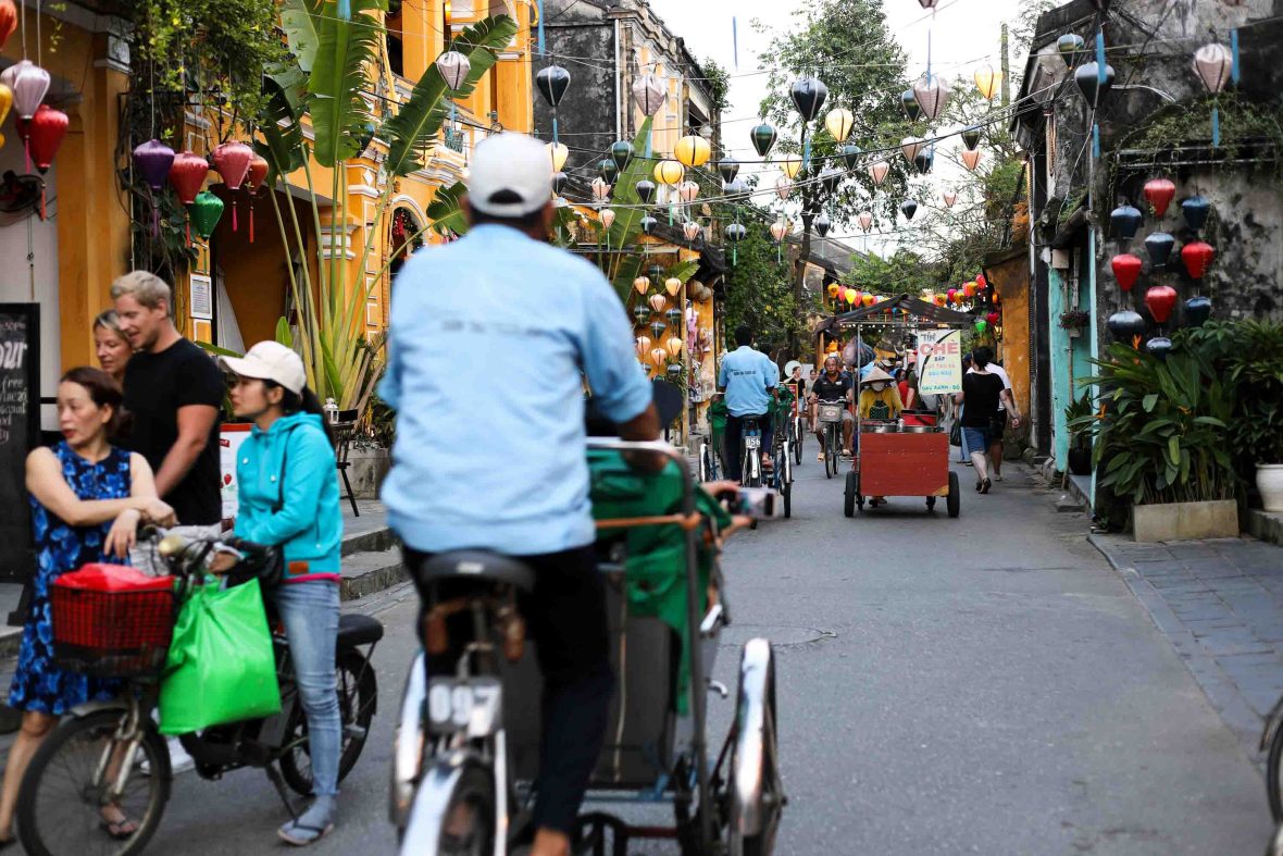 People walk and ride bikes through a narrow street with lanterns overhead.