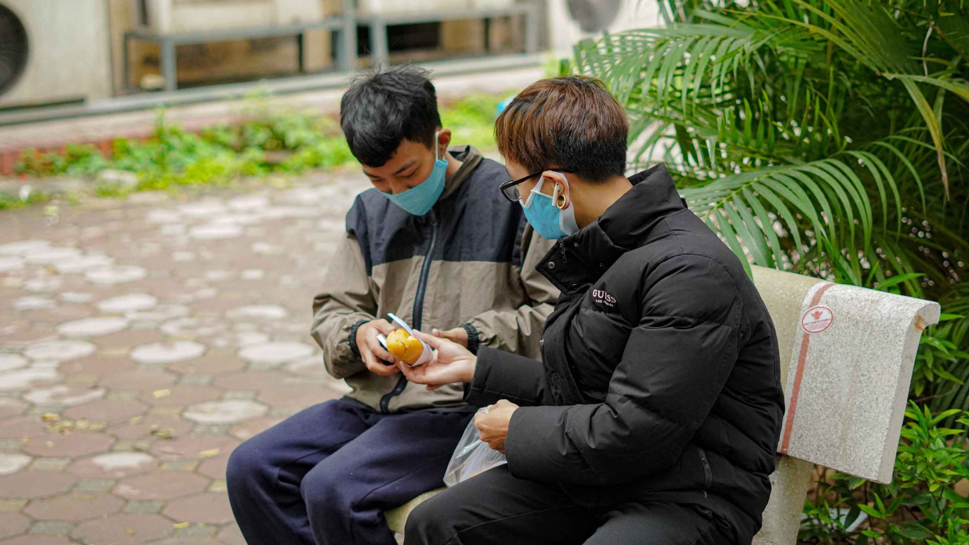 Two men sit together wearing face masks. One is handing the other a sandwich.